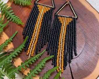 May include: A pair of long, black and gold beaded earrings with a triangular shape at the top. The earrings are hanging from gold hooks.