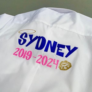 May include: White shirt with the name "SYDNEY" in blue glitter, a gold halo, and the years "2019-2024" in pink glitter. A gold glitter kiss mark is also present on the shirt.