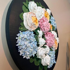 May include: A round, black wall art piece featuring a 3D floral arrangement. The flowers include white roses, pink and yellow peonies, and blue hydrangeas, with green leaves. The artwork is mounted on a black wooden base.