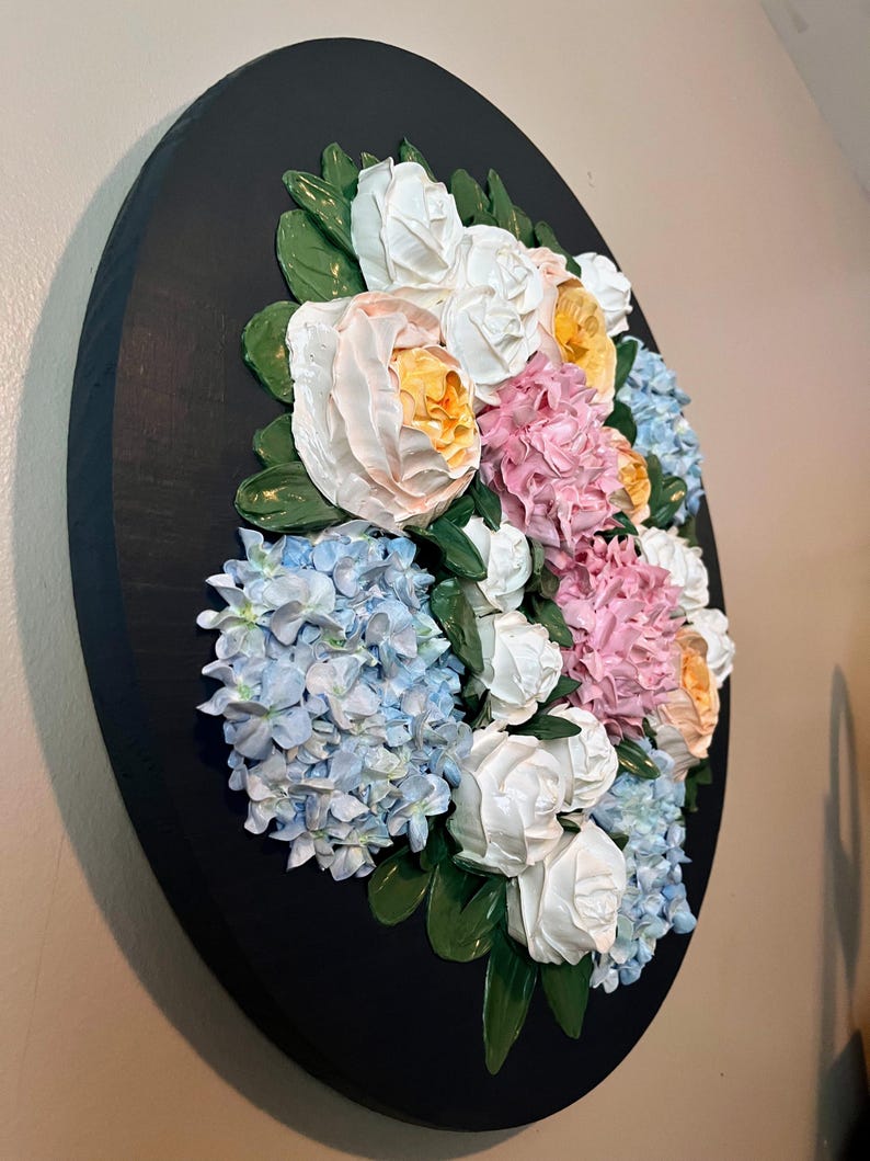 May include: A round, black wall art piece featuring a 3D floral arrangement. The flowers include white roses, pink and yellow peonies, and blue hydrangeas, with green leaves. The artwork is mounted on a black wooden base.