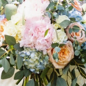 May include: A lush bouquet of flowers featuring a variety of blooms in soft pastel colors. The arrangement includes pink peonies, peach roses, blue hydrangeas, and white and yellow flowers, accented with green eucalyptus leaves.