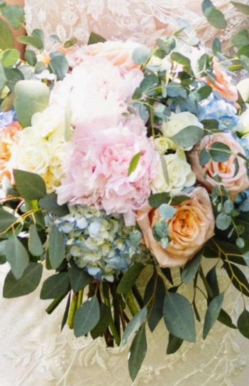 May include: A lush bouquet of flowers featuring a variety of blooms in soft pastel colors. The arrangement includes pink peonies, peach roses, blue hydrangeas, and white and yellow flowers, accented with green eucalyptus leaves.