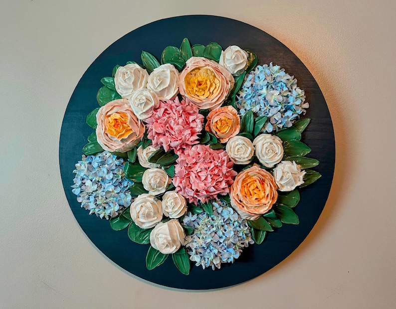 May include: A round, dark teal wooden wall hanging featuring a 3D floral arrangement. The arrangement includes pink, white, and orange roses, blue hydrangeas, and green leaves. The flowers are arranged in a circular pattern.