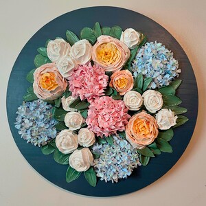 May include: A round, dark teal wall hanging featuring a floral arrangement. The arrangement includes pink hydrangeas, peach roses, and white roses, with green leaves. The flowers are arranged on a dark teal background.