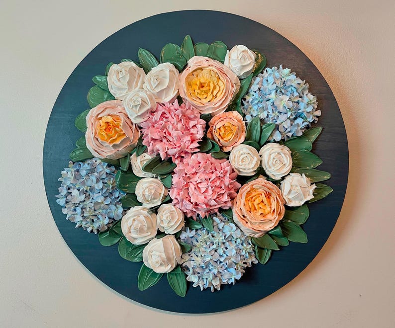 May include: A round, dark teal wall hanging featuring a floral arrangement. The arrangement includes pink hydrangeas, peach roses, and white roses, with green leaves. The flowers are arranged on a dark teal background.