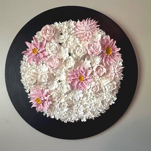 May include: A round, black wall art piece featuring a floral arrangement. The arrangement includes white roses and smaller white flowers, with pink dahlias and roses. The dahlias have yellow centers. The artwork is a decorative piece.