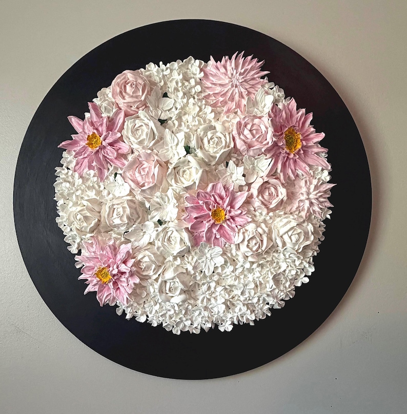 May include: A round, black wall art piece featuring a floral arrangement. The arrangement includes white roses and smaller white flowers, with pink dahlias and roses. The dahlias have yellow centers. The artwork is a decorative piece.