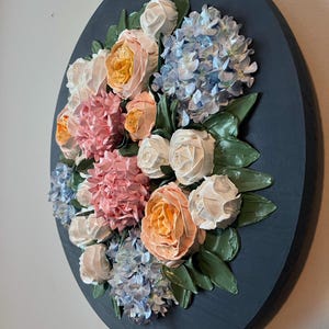 May include: A round, dark blue wall art piece featuring a textured floral arrangement. The flowers include peach, pink, white, and blue blooms with green leaves. The artwork is a 3D relief, creating a tactile and visually appealing piece.