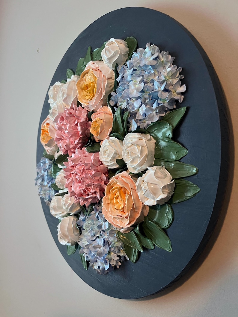 May include: A round, dark blue wall art piece featuring a textured floral arrangement. The flowers include peach, pink, white, and blue blooms with green leaves. The artwork is a 3D relief, creating a tactile and visually appealing piece.