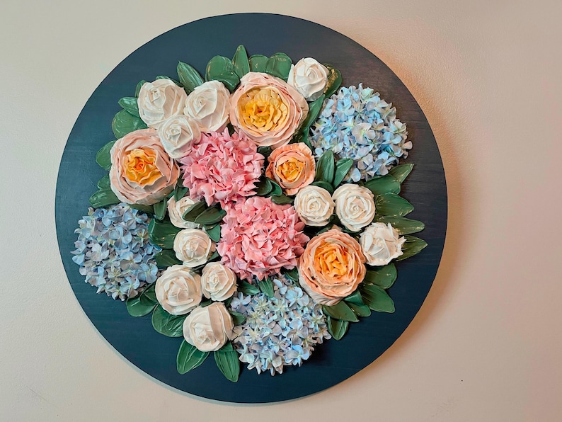 May include: A round, dark teal wall art piece featuring a floral arrangement. The arrangement includes pink, white, and peach-colored roses, blue hydrangeas, and green leaves. The flowers are arranged in a circular pattern.