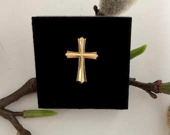 May include: A polished gold-tone cross pin featuring a sleek, multi-faceted design with sharp, tapered edges. The cross is centered on a square, black velvet-textured display card, creating a high-contrast look with its reflective metallic surface.