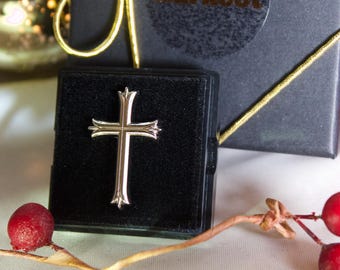 May include: A polished silver-toned metal cross pendant with flared ends, centered on a black velvet-lined square display box. The box is black with a gold ribbon and the text Karkoot visible on the lid, accompanied by red berries.