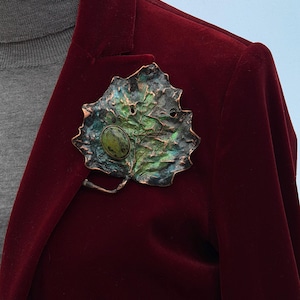 May include: A brooch shaped like a textured leaf with a copper-toned finish, featuring shades of green and black patina. An oval-shaped, mottled green stone is set on the left side of the leaf, which is pinned to a deep red velvet blazer.