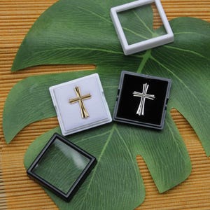 May include: Gold and silver cross pendants displayed in small square boxes, with a clear display case and a white frame. The crosses are set against white and black backgrounds, resting on a large green leaf.