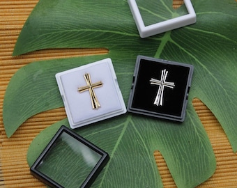 May include: Gold and silver cross pendants displayed in small square boxes, with a clear display case and a white frame. The crosses are set against white and black backgrounds, resting on a large green leaf.
