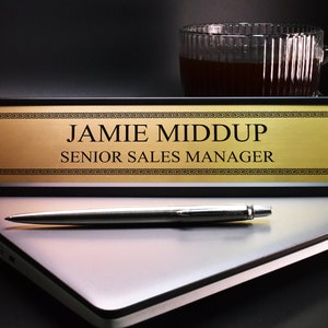 Könnte beinhalten: Ein goldenes Namensschild für den Schreibtisch mit schwarzem Rahmen und einem griechischen Schlüsselmuster. Das Namensschild lautet "Jamie Middup, Senior Sales Manager".