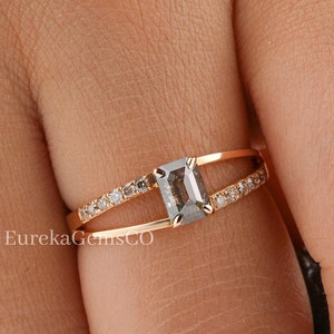 May include: A rose gold ring featuring a grey emerald-cut gemstone and smaller diamonds set in the band. The ring is worn on a person's finger.
