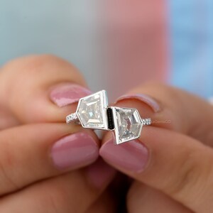 May include: A silver ring with two square-shaped diamonds set in a geometric design. The ring is set on a finger with pink nail polish.