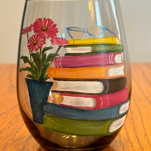 Pode incluir: Uma taça de vinho pintada à mão com uma pilha de livros, um par de óculos e uma planta em vaso com flores cor-de-rosa. Os livros são pintados em várias cores, incluindo vermelho, laranja, amarelo, verde e azul.