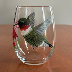 May include: A clear stemless wine glass with a hand-painted hummingbird design. The bird has green, red, and white feathers and outstretched wings. Red flowers are painted on the side. The glass is on a wooden surface.
