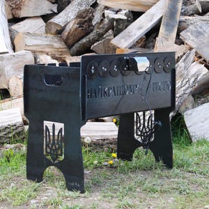 Può includere: Una griglia per barbecue in metallo nero con lo stemma ucraino ritagliato sulle gambe. La griglia ha una forma rettangolare con ritagli decorativi a forma di ingranaggio e testo sulla parte anteriore. La griglia è appoggiata sull'erba con un mucchio di legna da ardere sullo sfondo.