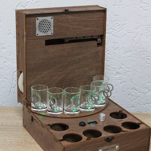 May include: A dark brown wooden box with a hinged lid, holding six small glass mugs. The box features a speaker, a control panel with a digital display, and a circular platform for the mugs. The box is stained.