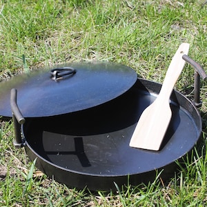 May include: A black metal fire pit with a lid and a wooden spatula. The fire pit is round and sits on the grass. The lid has a handle. The spatula is light-colored wood.