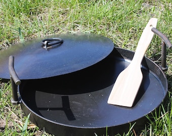 Peut inclure: Un foyer en métal noir avec un couvercle et une spatule en bois. Le foyer est rond et posé sur l'herbe. Le couvercle a une poignée. La spatule est en bois clair.