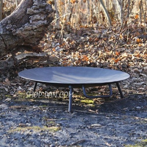 Puede incluir: Un brasero negro y redondo con patas cortas, colocado al aire libre. El brasero tiene forma de cuenco poco profundo y es de metal. La dirección del sitio web "SHOP-DAN.COM" está impresa en blanco en el suelo.