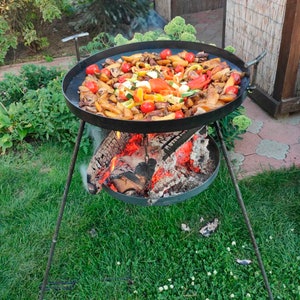 Pode incluir: Uma grande panela redonda de metal preto cheia de vegetais cozidos, incluindo batatas, tomates e pimentões, está sobre uma fogueira. A panela é suportada por um suporte de metal com três pernas, colocado em um gramado verde.
