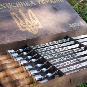 Puede incluir: Un juego de caja de madera con brochetas, con un emblema dorado y texto en ucraniano. La caja está abierta, revelando las brochetas con mangos de madera y varillas de metal. El texto dice "2023 P."