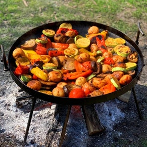 Puede incluir: Una gran sartén redonda de metal negro llena de verduras a la parrilla. Las verduras incluyen pimientos rojos y amarillos, calabacín, berenjena y patatas. La sartén está sostenida por un trípode sobre un lecho de brasas.