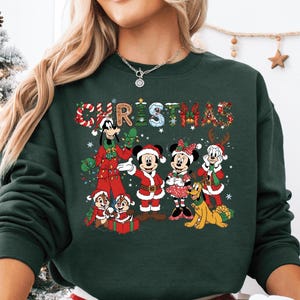 May include: Dark green Christmas sweatshirt featuring Mickey Mouse, Minnie Mouse, and other Disney characters in festive outfits. The word "CHRISTMAS" is written across the top in colorful letters. The person is wearing red and white plaid pajama pants and holding a mug with Santa Claus.