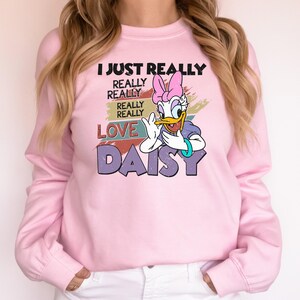 May include: A light pink sweatshirt featuring Daisy Duck from Disney. The graphic includes the text "I JUST REALLY REALLY REALLY LOVE DAISY" with Daisy Duck in a purple top and pink bow. The sweatshirt has long sleeves and a crew neck.