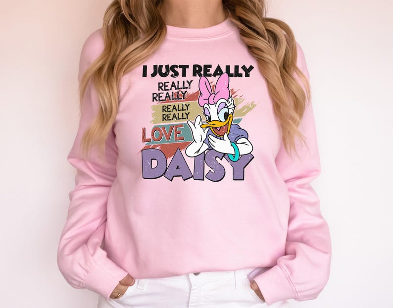 May include: A light pink sweatshirt featuring Daisy Duck from Disney. The graphic includes the text "I JUST REALLY REALLY REALLY LOVE DAISY" with Daisy Duck in a purple top and pink bow. The sweatshirt has long sleeves and a crew neck.
