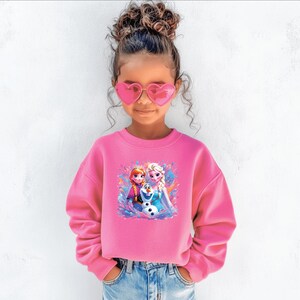 May include: A young girl wearing a bright pink sweatshirt with a colorful graphic of animated characters. The sweatshirt features a crew neck and long sleeves. She is also wearing heart-shaped pink sunglasses and blue jeans.