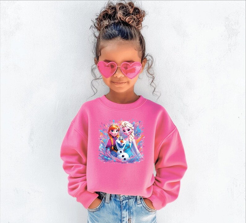 May include: A young girl wearing a bright pink sweatshirt with a colorful graphic of animated characters. The sweatshirt features a crew neck and long sleeves. She is also wearing heart-shaped pink sunglasses and blue jeans.