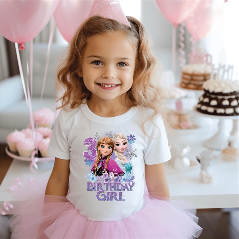 May include: A young girl wearing a white t-shirt with a colourful graphic featuring the number 3, and the words "Birthday Girl" with images of two animated characters. She is also wearing a pink tutu and a party hat.