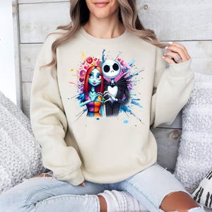 Op de afbeelding: Beige sweatshirt met een aquarelontwerp van Jack Skellington en Sally uit The Nightmare Before Christmas. De afbeelding toont de personages in een hartvorm met blauwe, roze en gele verfspatten.