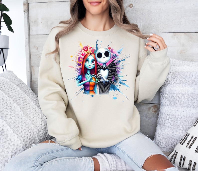 Op de afbeelding: Beige sweatshirt met een aquarelontwerp van Jack Skellington en Sally uit The Nightmare Before Christmas. De afbeelding toont de personages in een hartvorm met blauwe, roze en gele verfspatten.