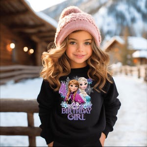 May include: A young girl wearing a black sweatshirt and a pink knit hat. The sweatshirt features a graphic of two animated characters, the number 4, and the words "BIRTHDAY GIRL" in purple.