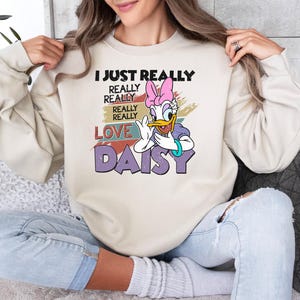 May include: Beige sweatshirt featuring a graphic of Daisy Duck with a pink bow and the text "I JUST REALLY REALLY REALLY LOVE DAISY". The design has a vintage, distressed look. The sweatshirt is a casual, comfortable style.