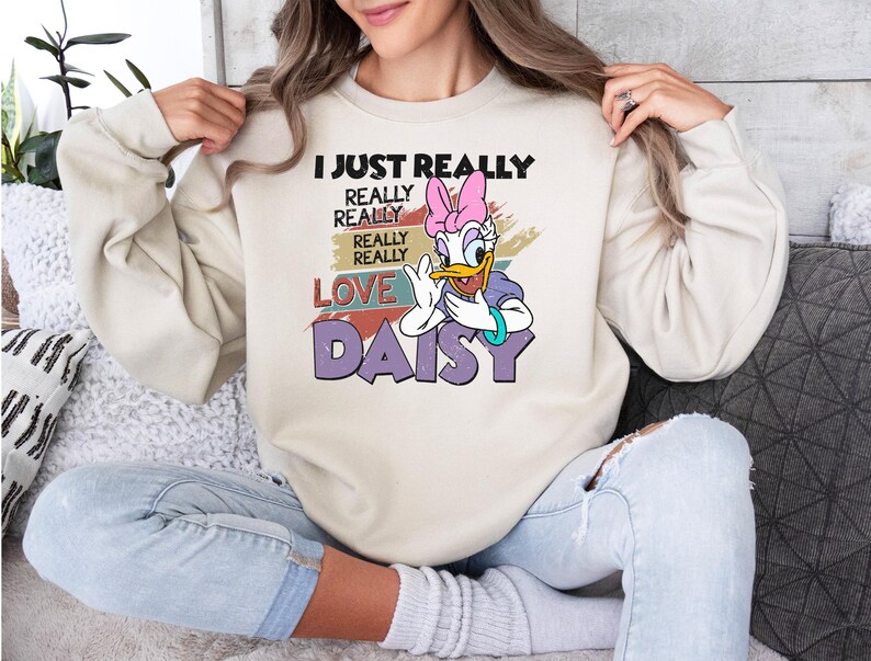 May include: Beige sweatshirt featuring a graphic of Daisy Duck with a pink bow and the text "I JUST REALLY REALLY REALLY LOVE DAISY". The design has a vintage, distressed look. The sweatshirt is a casual, comfortable style.