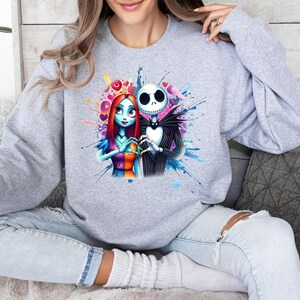 Op de afbeelding: Een lichtgrijs sweatshirt met een kleurrijke afbeelding van Jack Skellington en Sally uit The Nightmare Before Christmas. De illustratie toont de personages in een hartvorm met een aquareleffect. De sweatshirt heeft een ronde hals.