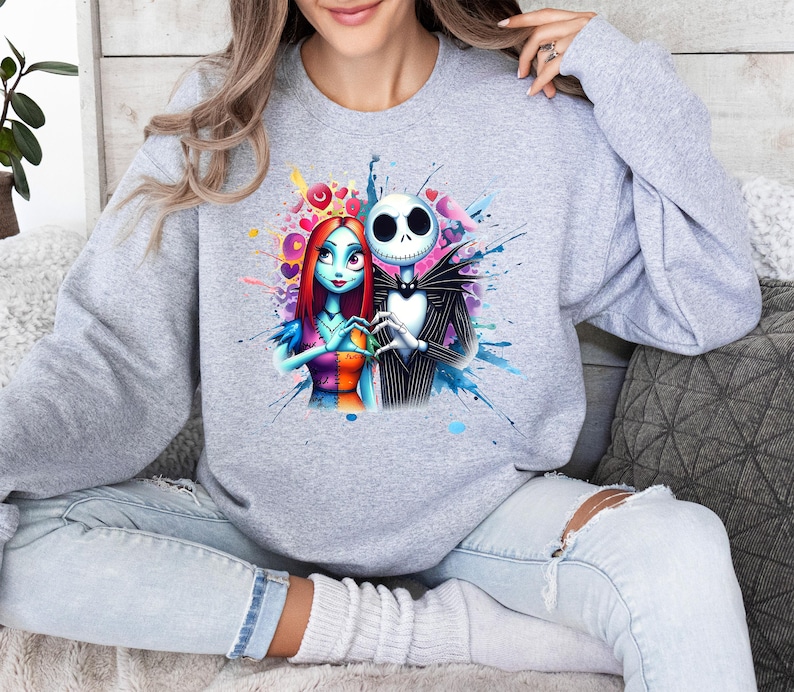 Op de afbeelding: Een lichtgrijs sweatshirt met een kleurrijke afbeelding van Jack Skellington en Sally uit The Nightmare Before Christmas. De illustratie toont de personages in een hartvorm met een aquareleffect. De sweatshirt heeft een ronde hals.