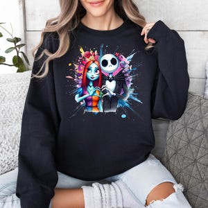 Op de afbeelding: Zwart sweatshirt met een kleurrijk aquarelontwerp van Jack Skellington en Sally uit The Nightmare Before Christmas. De afbeelding toont de personages die elkaars hand vasthouden, omringd door levendige kleurspatten.