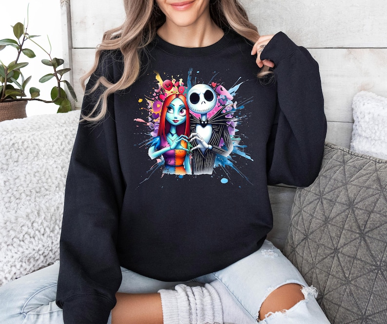 Op de afbeelding: Zwart sweatshirt met een kleurrijk aquarelontwerp van Jack Skellington en Sally uit The Nightmare Before Christmas. De afbeelding toont de personages die elkaars hand vasthouden, omringd door levendige kleurspatten.