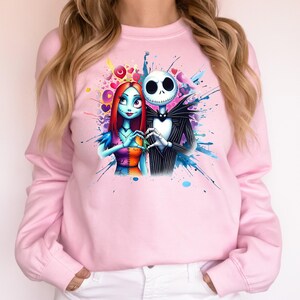 Op de afbeelding: Lichtroze sweatshirt met een kleurrijke afbeelding van Jack Skellington en Sally uit The Nightmare Before Christmas. De personages vormen een hart met hun handen, omringd door kleurrijke spetters.