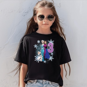 May include: Black t-shirt featuring a graphic of Elsa, Anna, and Olaf from Frozen, with the number 3 in glitter. The design is surrounded by snowflakes. The shirt is worn by a young girl wearing heart-shaped sunglasses.