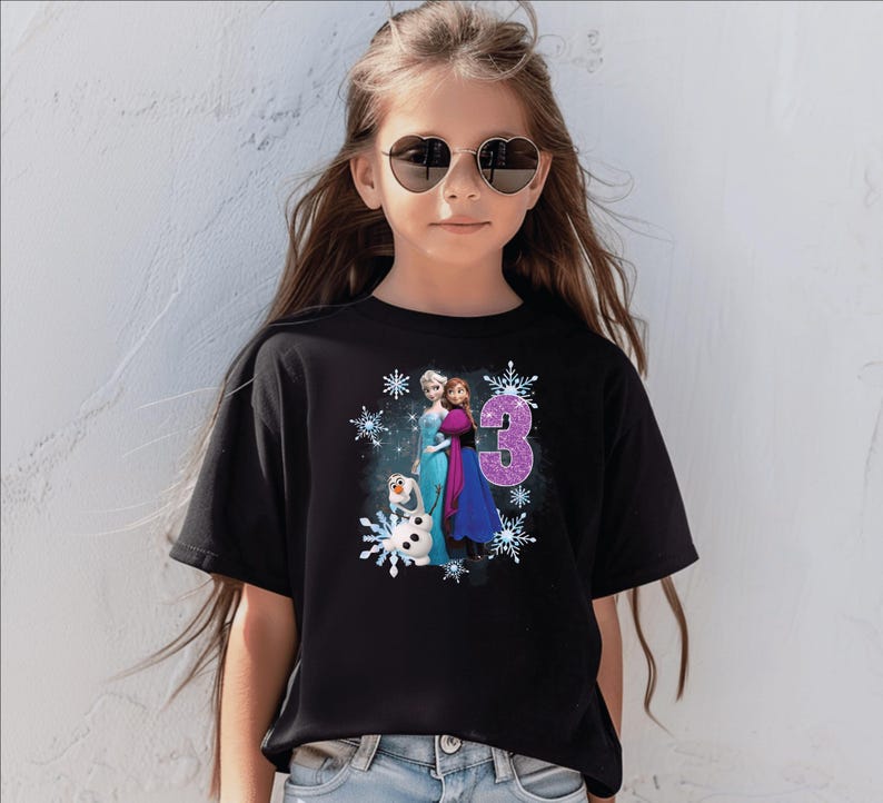 May include: Black t-shirt featuring a graphic of Elsa, Anna, and Olaf from Frozen, with the number 3 in glitter. The design is surrounded by snowflakes. The shirt is worn by a young girl wearing heart-shaped sunglasses.